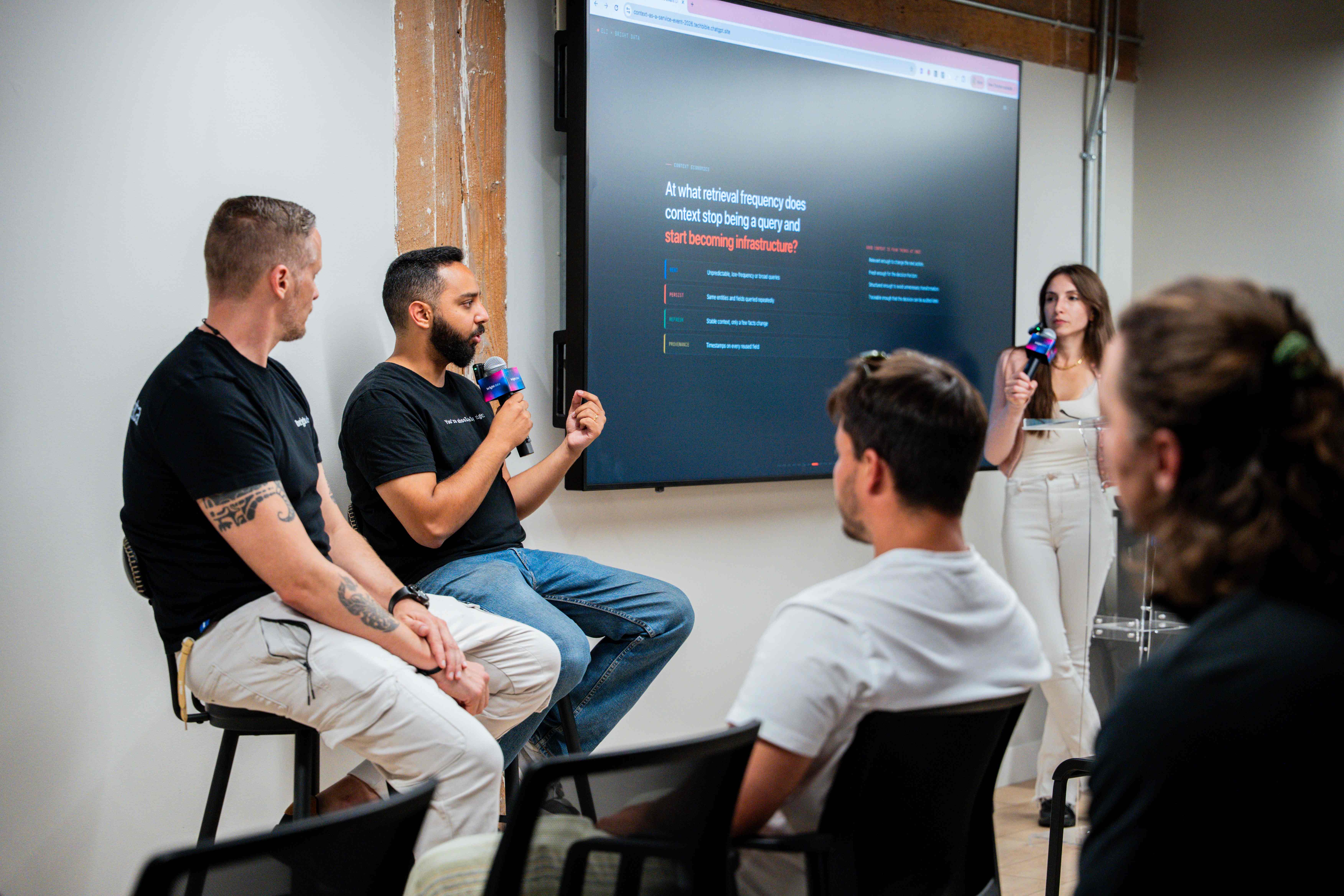 Panel discussion at an H2ai event, two speakers on stools with a host and a seated audience, a slide behind them asking when context stops being a query and starts becoming infrastructure.