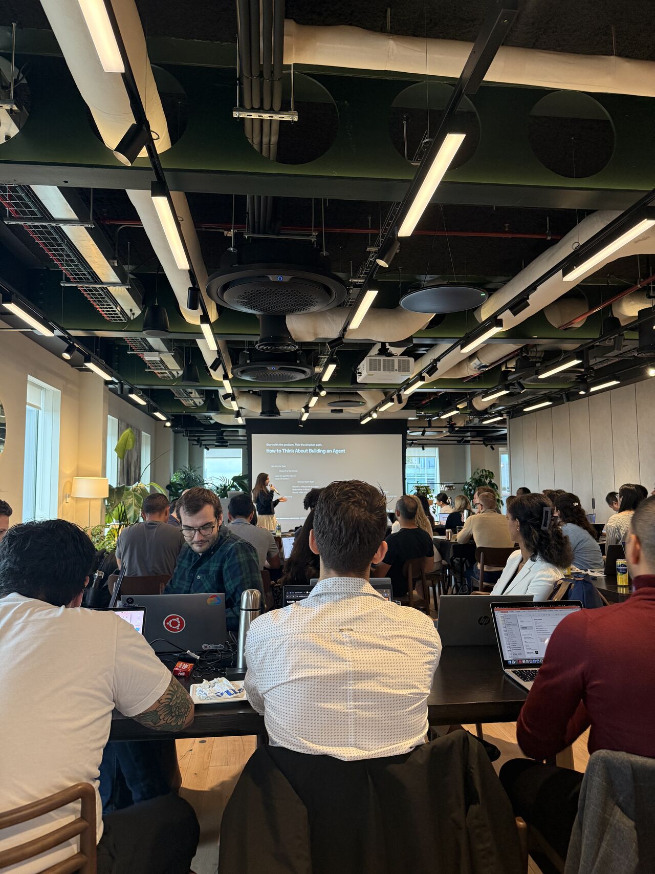 A full room of builders at long tables with laptops during the Dawn Capital hackathon, a presenter at the front beside a screen reading How to Think About Building an Agent.