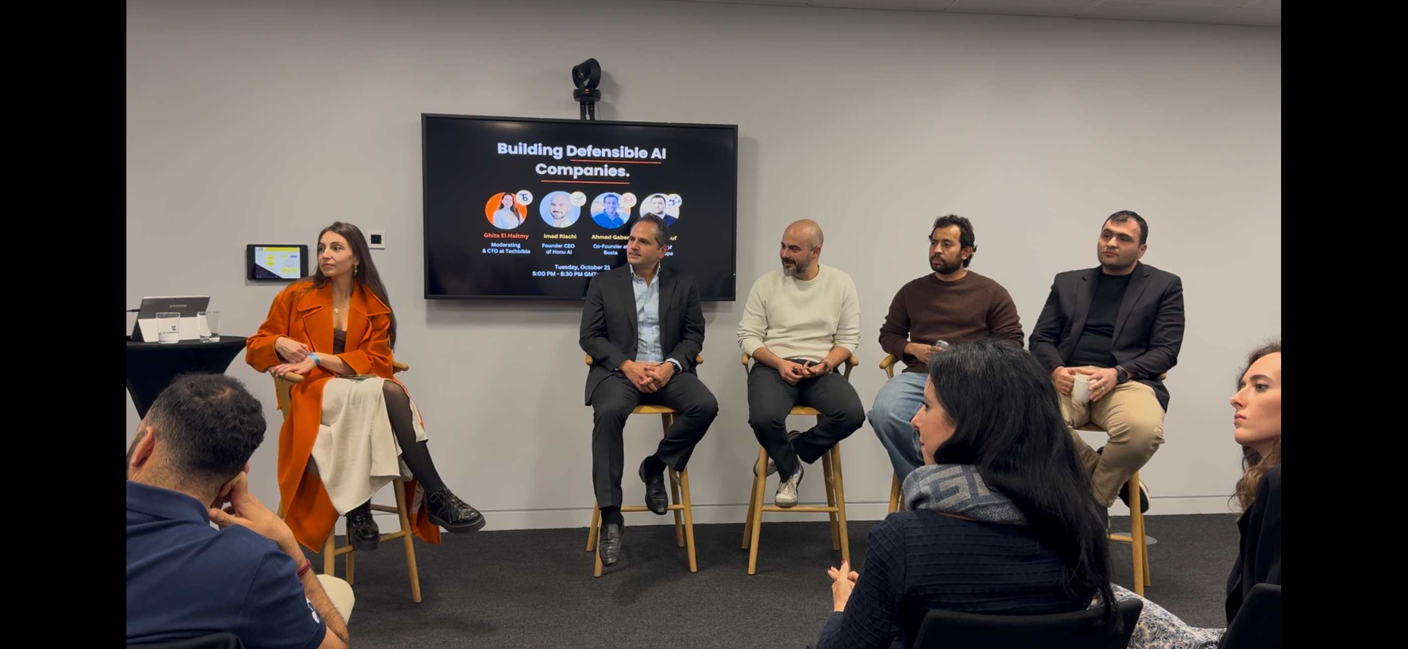 Ghita moderating the Building Defensible AI Companies panel at One Canada Square, seated beside three panelists on stools in front of a screen showing the event title and speaker line-up.