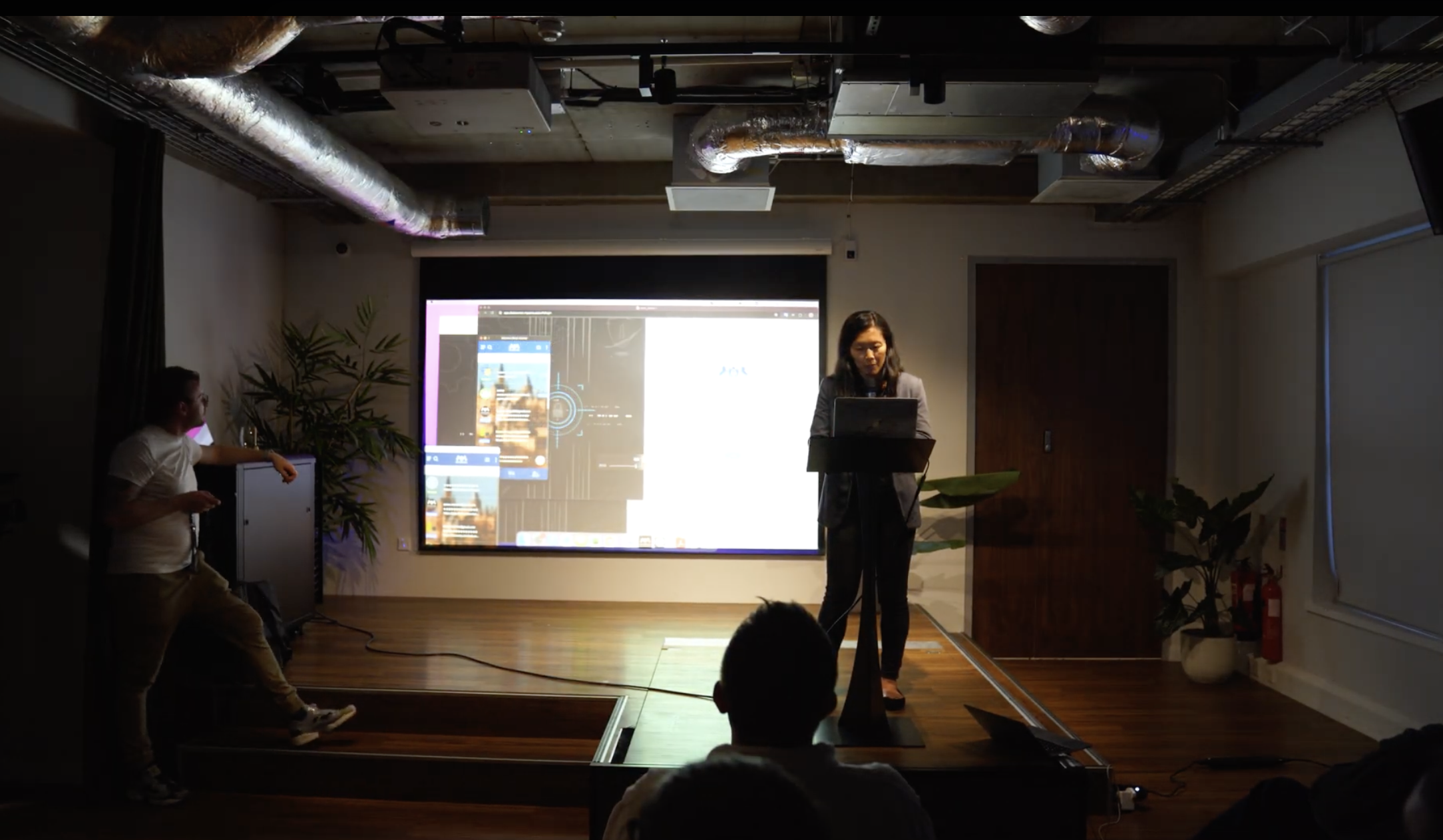 A founder presenting from a podium on a dimly lit stage at AI Startup Roast Night, her product demo projected on the screen behind her as the audience watches.